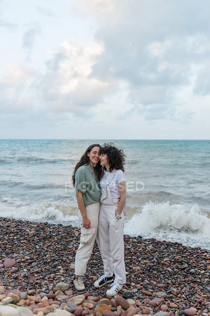 Jóvenes novias lesbianas en ropa casual abrazándose mientras se miran en la costa del océano bajo el cielo nublado - foto de stock