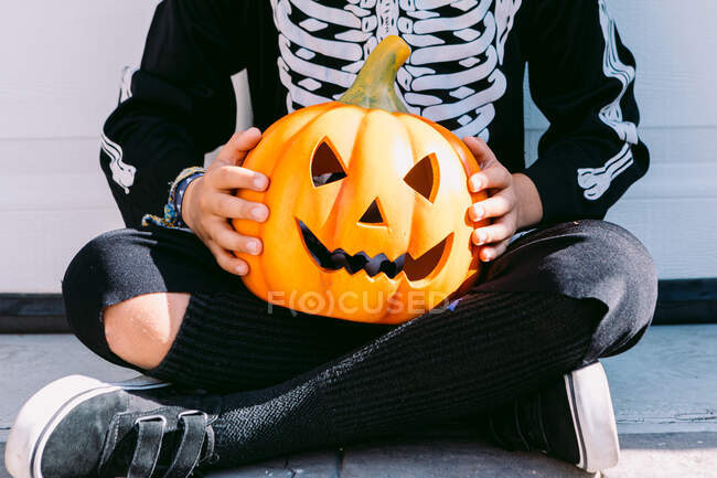Crop unrecognizable child in black skeleton costume holding creepy carved Halloween Jack O Lantern pumpkin while sitting with legs crossed on street — Stock Photo