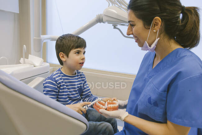 Orthodontiste féminine souriante enseignant au patient avec une brosse à dents à brosser les dents sur un modèle de mâchoire dans une clinique dentaire — Photo de stock