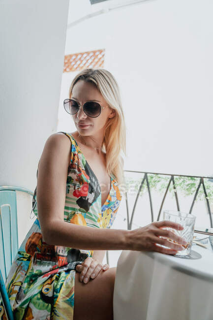 Вид збоку молодої жінки в сонцезахисних окулярах, що сидить зі склянкою віскі за столом з подругою врожаю на балконі — стокове фото