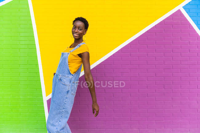 Vista lateral de la hembra afroamericana feliz sonriendo mientras salta por encima del suelo cerca de la pared brillante - foto de stock