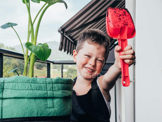 Niño positivo con pala de jardinería sucia mirando cámara contra bolsa con planta tropical en el campo - foto de stock