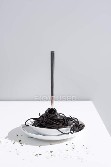 Stainless fork in full bowl of black spaghetti with cuttlefish ink on white table with thin blooms in studio on gray background — Stock Photo