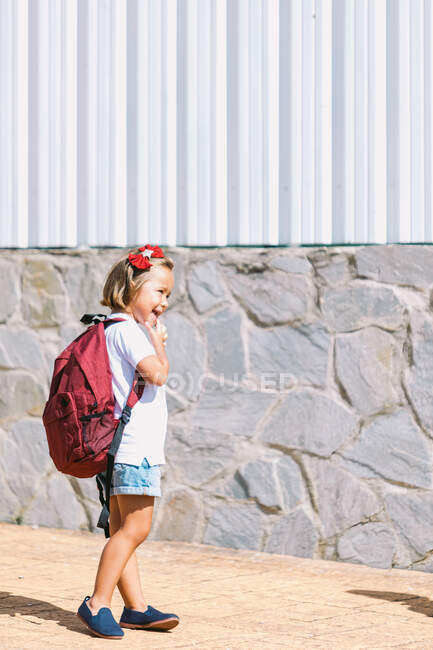 Вид збоку школяра з рюкзаком, який торкається губ на тротуарі, дивлячись вперед на сонячне світло — стокове фото
