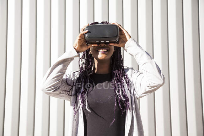 Jovem afro-americana excitada no fone de ouvido VR entretendo e jogando jogo virtual contra a parede listrada cinza — Fotografia de Stock