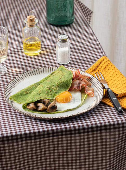 Pequeno-almoço caseiro de panquecas de espinafre com bacon, ovo e cogumelos servido em uma placa branca com um sal e óleo agitador em uma toalha de mesa quadriculada. — Fotografia de Stock