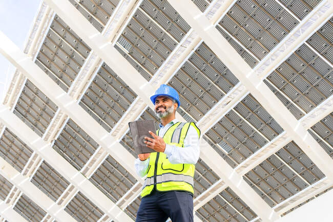 Низький кут щасливого середнього віку інженер - латиноамериканець у формі з планшетом, що стоїть осторонь під сонячною електростанцією. — стокове фото