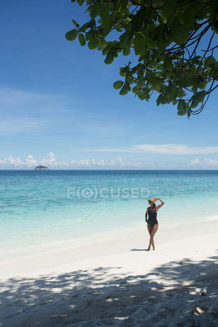 Этническая туристка в купальнике и соломенной шляпе, идущая по песку во время поездки в Малайзию — стоковое фото