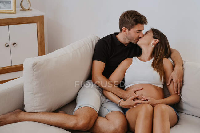 Homem beijando e abraçando barriga de expectante amado feminino enquanto descansa no sofá na sala de estar — Fotografia de Stock