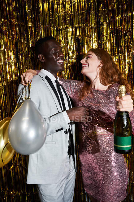 Приємна молода жінка з пляшкою шампанського і кулями, які обіймають афроамериканського чоловіка, коли він дивиться один на одного під час вечірки. — стокове фото