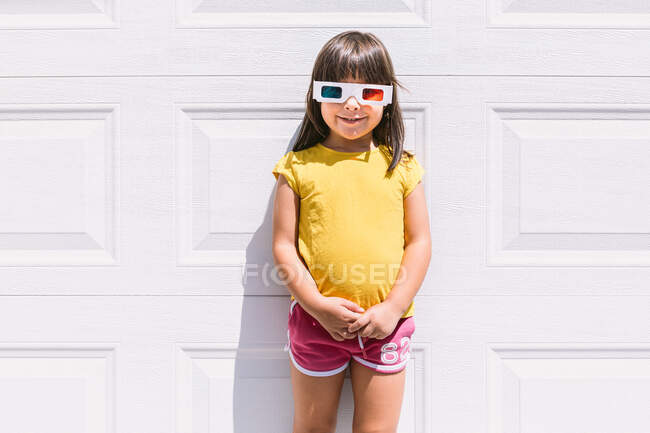 Joyeux fille mignonne dans des vêtements colorés occasionnels et des lunettes tridimensionnelles debout sur fond de mur blanc — Photo de stock