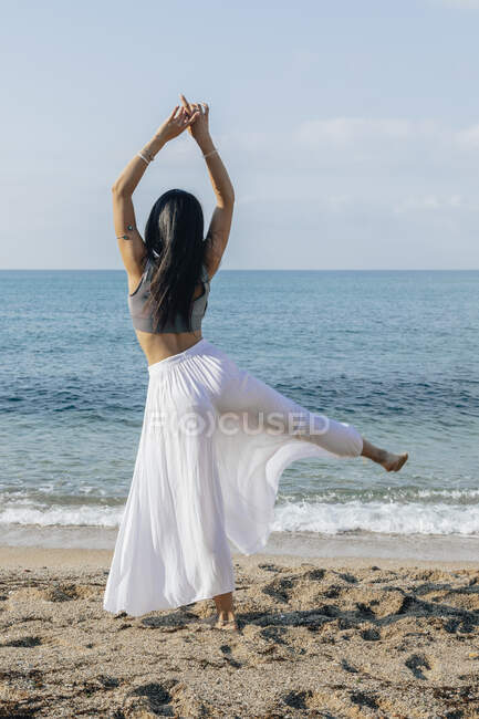 Visão traseira da fêmea anônima com braços levantados durante a prática de ioga na costa verde contra o oceano — Fotografia de Stock