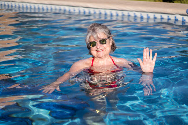 Cheerful senior female in bikini inside of poolside in clean water and waving — Stock Photo