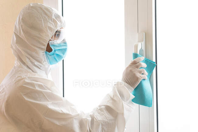 Side view of male medic in personal protective equipment cleaning handle of window with rag during COVID 19 pandemic in hospital — Stock Photo