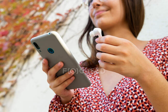 Знизу врожаю весела жінка в повсякденному одязі, що з'єднує бездротові навушники зі смартфоном проти світлої стіни вдень — стокове фото