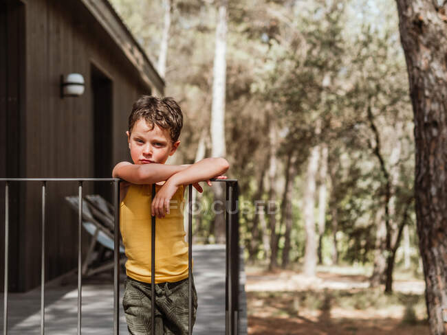 Menino de pé na varanda da casa de campo moderna localizada na floresta no verão — Fotografia de Stock