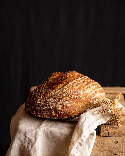 Delicioso pan artesanal entero con corteza sobre tela arrugada contra espigas de trigo en superficie de madera - foto de stock
