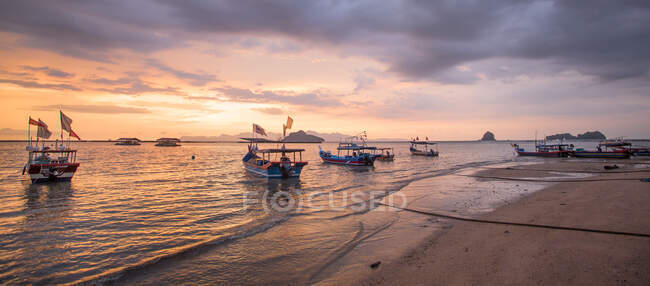 Широкий угол живописного вида деревянных лодок на волнистом море, омывающих песчаное побережье под облачным небом на закате солнца в Малайзии — стоковое фото