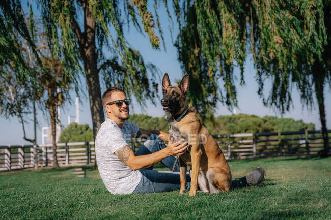 Side view of tattooed male in sunglasses with Malinois resting on meadow and looking at each other — Stock Photo