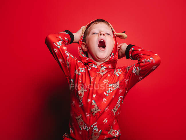 Menino engraçado em pijama de Natal com capuz com ornamento de veado bocejando e olhando para a câmera contra o fundo vermelho — Fotografia de Stock