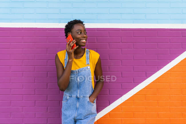 Задоволена афро-американська жінка стоїть з рукою в кишені біля яскравої стіни і розмовляє по мобільному телефону, дивлячись на — стокове фото