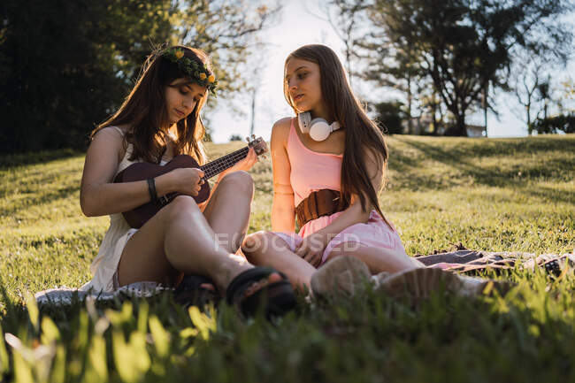 Adolescente em coroa de flores jogando ukulele enquanto se senta contra a melhor amiga com fones de ouvido sem fio na parte traseira iluminada — Fotografia de Stock