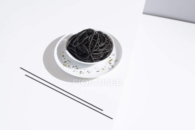 Сверху керамической миски с вкусными спагетти с черными чернилами кальмара с палочками на белом фоне — стоковое фото