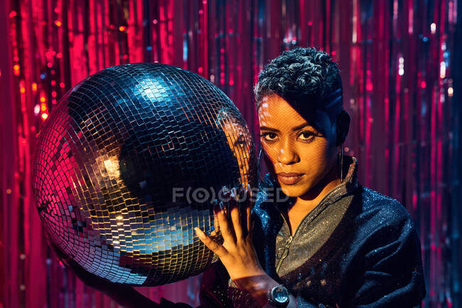 Menina étnica jovem na moda olhando para a câmera enquanto segurava bola espelho na boate — Fotografia de Stock