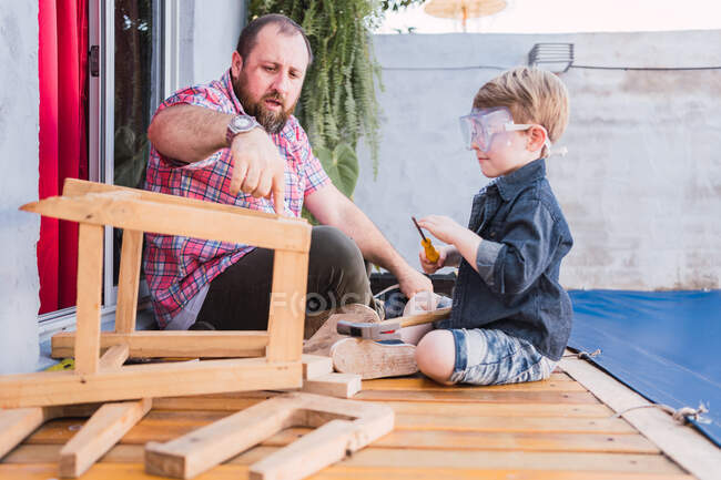 Mature dad explaining to son in protective glasses how to drive screw into wooden block while sitting on boardwalk — Stock Photo