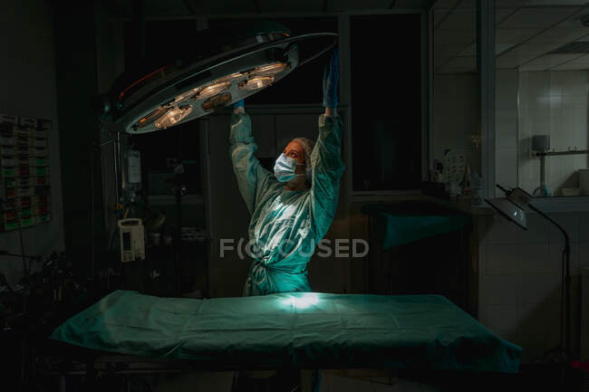 Молода жінка-ветеринар у хірургічній уніформі та стерильній масці, дивлячись вгору під час коригування лампи над медичним текстилем у клініці — стокове фото