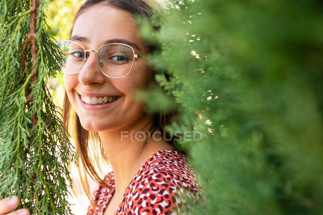 Cheerful young female with brown hair in eyeglasses standing among green branches and looking at camera in daylight — Stock Photo