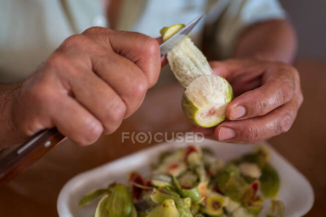 Обрізаний невпізнаваний старший чоловік з ножем чистить зелений інжир за столом з одноразовим лотком в домашній кімнаті — стокове фото