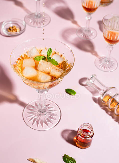 From above of glass with whiskey and ice cubes garnished with basil leaves served on pink table with shiny goblets — Stock Photo