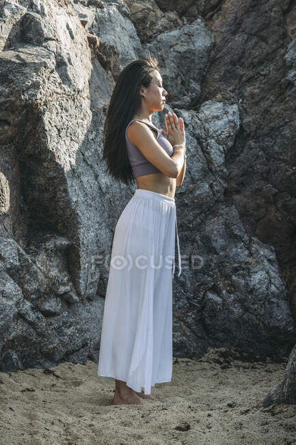 Боковой вид этнической женщины с молящимися руками, практикующей йогу на песчаной земле против скалы в солнечном свете — стоковое фото