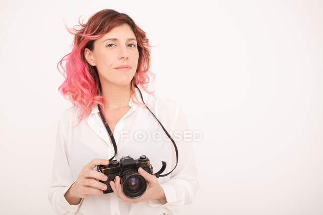 Fotógrafa pensativa con el pelo rosa sosteniendo una cámara fotográfica profesional en sus manos en la sala de luz - foto de stock