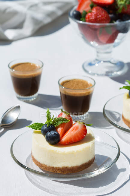 Tasty cheesecakes with fresh strawberries and blueberries under mint leaves against shots of espresso coffee on table in cafeteria — Stock Photo