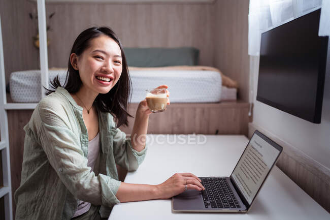 Vista lateral del contenido joven blogger étnica en el escritorio con netbook y café mirando a la cámara en la habitación de la casa - foto de stock