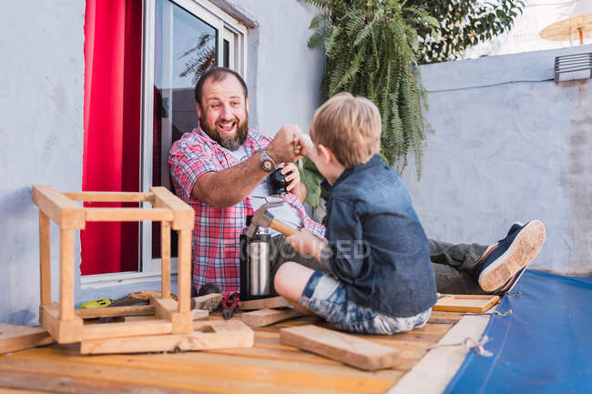 Ground level of cheerful bearded dad in checkered shirt against boy bumping fists while looking at each other against wooden blocks — Stock Photo