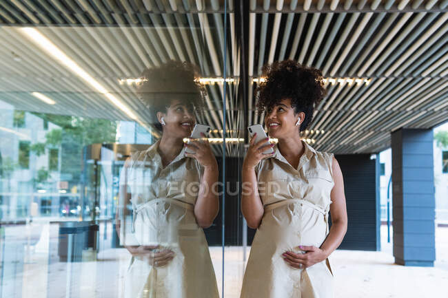 Pleasant pregnant African American female in wireless earbuds with curly hair recording voice message on cellphone near glass building — Stock Photo