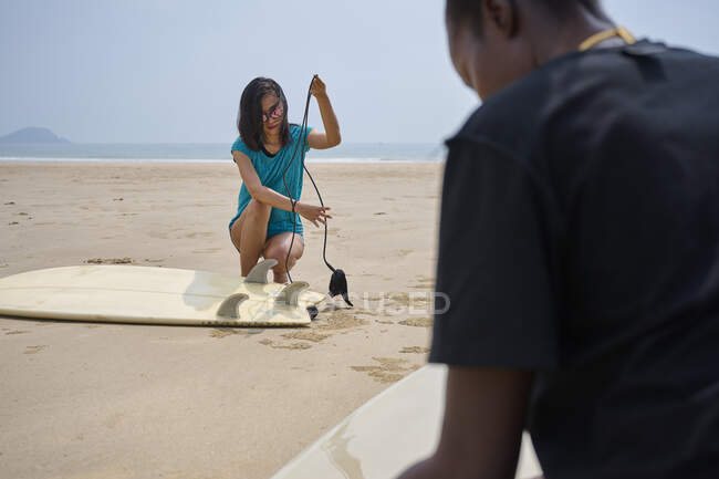 Jovem esportista asiática com caixa de barbatana em corda agachada contra prancha de surf e colheita anônima namorada negra na costa — Fotografia de Stock