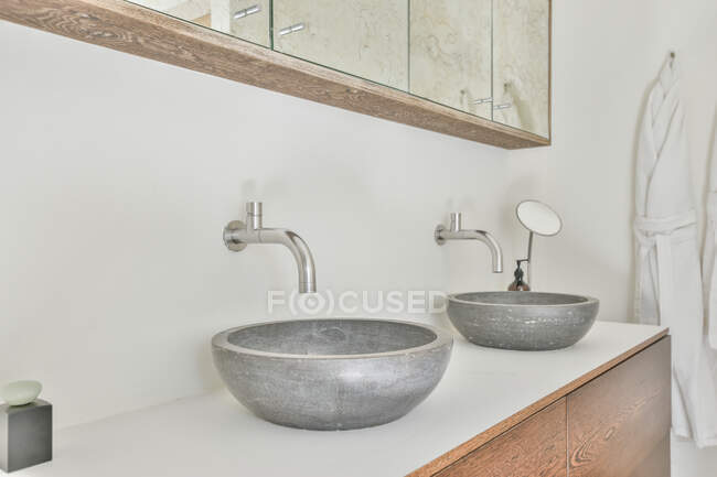 Lavabos redondos con grifos entre armarios con espejos contra albornoz en casa ligera - foto de stock