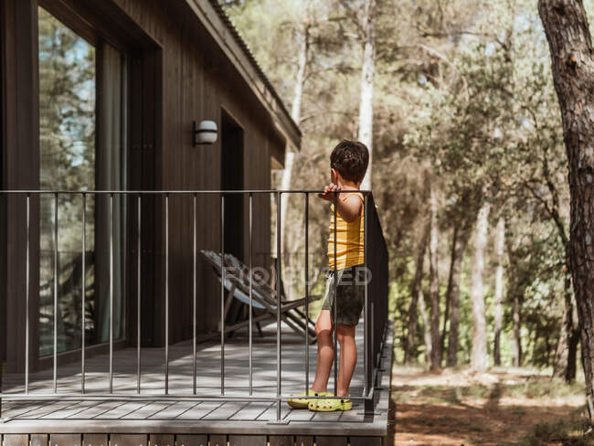 Вид збоку на невпізнаваного маленького хлопчика, що стоїть на веранді сучасного котеджу, розташованого в лісі влітку — стокове фото