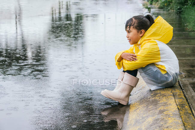 Seitenansicht eines verträumten ethnischen Kindes in Regenmantel und Gummistiefeln, das mit verschränkten Armen auf dem Bürgersteig sitzt und sich auf Regentage freut — Stockfoto