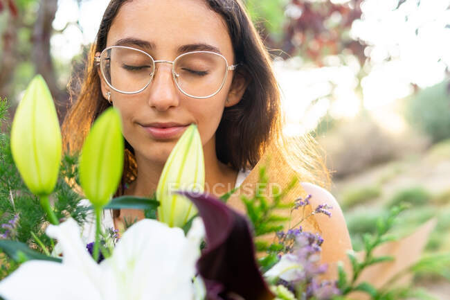 Cosecha joven mujer consciente con los ojos cerrados en gafas disfrutando del aroma del floreciente ramo floral en la ciudad sobre un fondo borroso - foto de stock