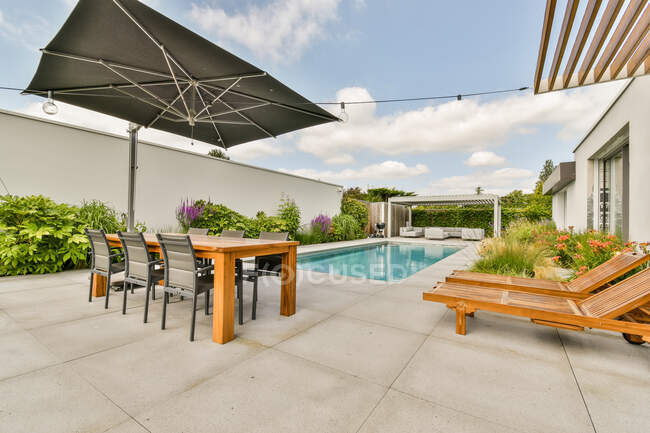 Esstisch mit Stuhl und hölzernen Liegestühlen in der Nähe des Swimmingpools im Hof einer teuren zeitgenössischen minimalistischen Villa an sonnigen Tagen — Stockfoto