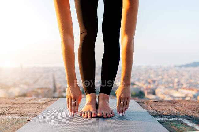 Crop unrecognizable barefoot female in leggings performing Uttanasana pose while practicing yoga on mat on rooftop — Stock Photo