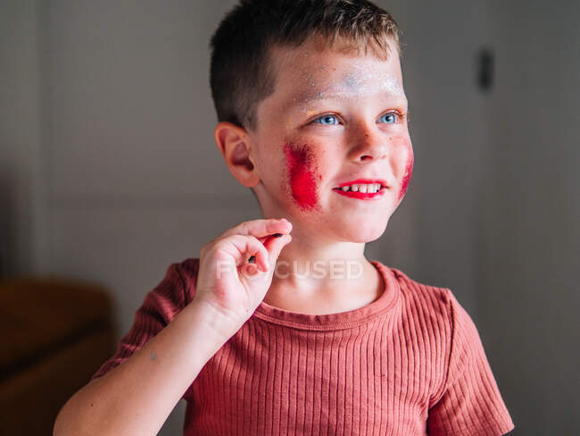 Niño desordenado con cabello castaño y cosmético en la cara en casa de luz sobre fondo borroso - foto de stock
