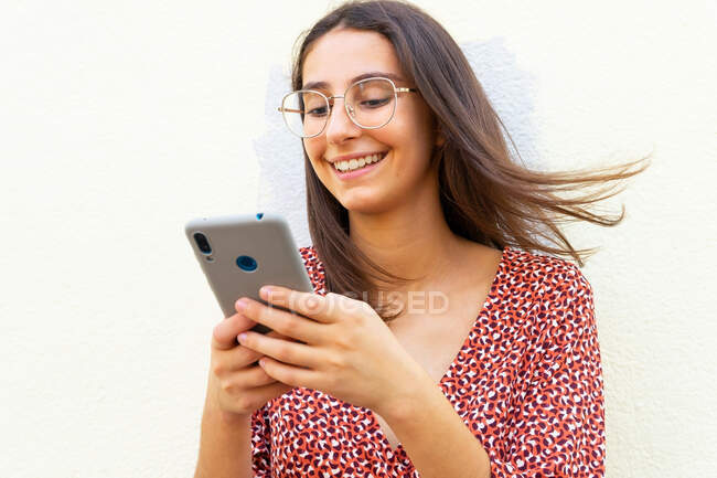 Улыбающаяся женщина в платье и очках стоит возле белой стены и днем пользуется смартфоном — стоковое фото