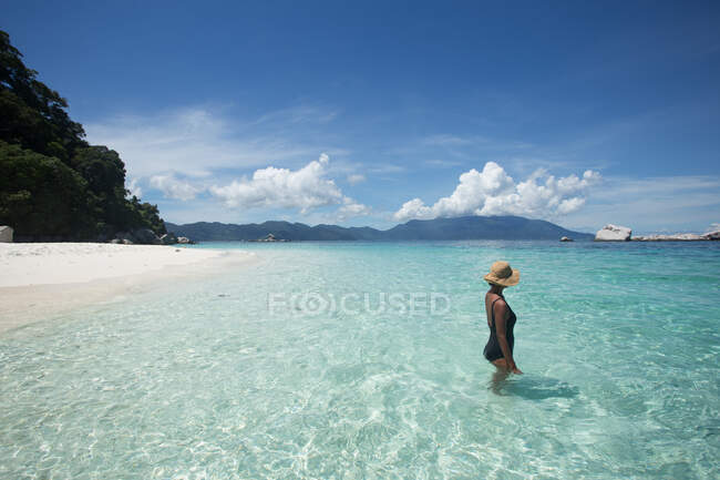 Vista lateral de turista femenina étnica en traje de baño y sombrero de paja de pie en el mar transparente durante el viaje en Malasia - foto de stock