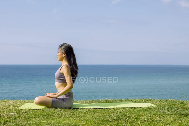 Vista lateral de la joven étnica femenina en ropa deportiva meditando con las piernas cruzadas durante la práctica de yoga contra el mar - foto de stock